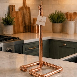 May include: A vertical paper towel holder constructed from polished copper pipes and fittings. The base is square-shaped with a central upright rod featuring a small hook at the top. A white square tag with the text THE COPPER LINE is attached.