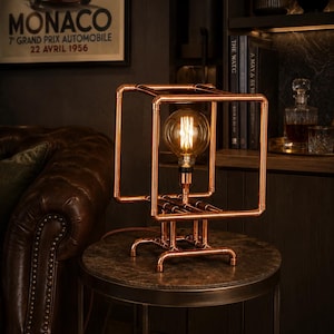 Op de afbeelding: Een tafellamp gemaakt van koperkleurige metalen buizen in een kubusvormig frame. De lamp heeft een centrale fitting met een ronde, amberkleurige Edison-stijl gloeilamp met zichtbare filamenten, rustend op een buisvoet.