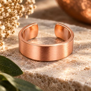 May include: A copper-coloured open band ring with a brushed metal finish. The ring features a simple, wide, flat design with a gap at the top, showcasing a warm metallic tone and a textured surface that reflects light across its circular form.