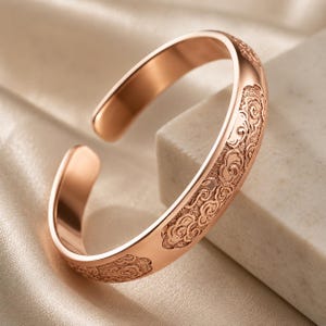 May include: A rose gold cuff bracelet featuring an open design and a polished finish. The outer surface is decorated with a repeating embossed pattern of stylized swirling clouds, providing a textured contrast to the smooth metal edges.