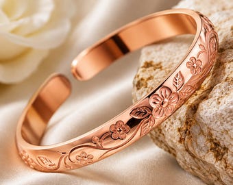 May include: A rose gold-toned open cuff bracelet featuring an embossed floral pattern with delicate flowers and leaves winding along the outer surface. The metal has a polished, reflective finish that catches the light, highlighting the intricate botanical engravings.