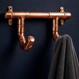 May include: A wall-mounted towel hook rack constructed from polished copper piping and fittings. The horizontal bar features two curved hooks extending downward, with a dark grey textured towel draped over the right hook and gold-toned screws.