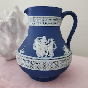 May include: A blue ceramic pitcher featuring white relief decorations. The design includes a central frieze of three dancing figures and two horizontal bands of intricate floral lattice patterns. It has a curved handle and a pouring spout.