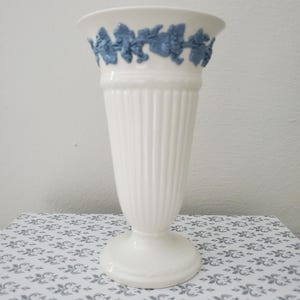 May include: A white ceramic vase with a flared rim and a vertical ribbed body. The upper section features a raised, light blue relief decoration depicting grapevines and leaves encircling the neck. The vase stands on a circular pedestal base.