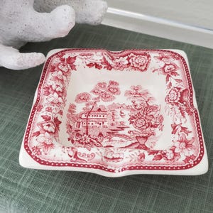 May include: A square ceramic ashtray featuring a white base with a detailed red transferware pattern. The rim displays a floral border, while the center depicts a landscape scene with a building, trees, and a bridge. It has four indentations.