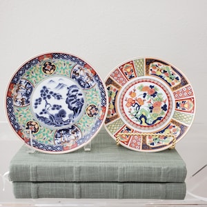 May include: Two decorative ceramic plates featuring intricate patterns. The left plate displays a blue and white landscape with a pine tree. The right plate features a colorful floral design with chrysanthemums and geometric borders.
