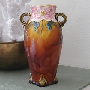 May include: A ceramic vase featuring a glossy amber and brown body with two side handles. The rim is decorated with a sculpted pink flower and green leaf accents. The base has a scalloped edge, and the overall finish is smooth and reflective.