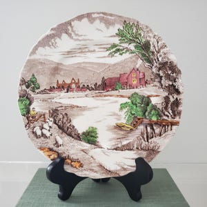 May include: A round ceramic plate with a scalloped edge featuring a brown and white transferware landscape scene. The image depicts a river, a red building, green trees, and a person tending to a small flock of white sheep near a wooden fence.