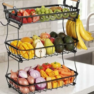 May include: A three-tier black metal wire storage rack with wooden handles, filled with various fresh produce including strawberries, grapes, kiwi, bananas, pears, apples, mangoes, red onions, tomatoes, bell peppers, and carrots in separate baskets.