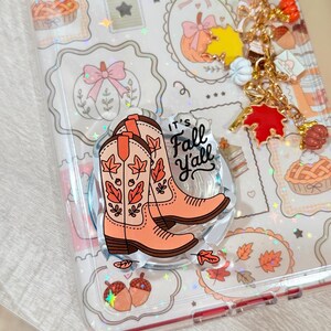 May include: A clear phone case featuring a holographic autumn pattern of pies, pumpkins, and acorns. An acrylic phone grip displays two orange cowboy boots and the text "It's Fall Y'all." A gold-toned charm chain with miniature pumpkin and leaf pendants is attached.