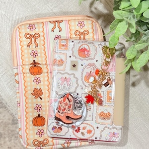 May include: A rectangular tablet case with a peach and white gingham pattern featuring orange pumpkins, brown leaves, and pink bows. A clear tablet cover sits on top, decorated with fall-themed stickers, a cowboy boot pop-socket, and a gold charm.