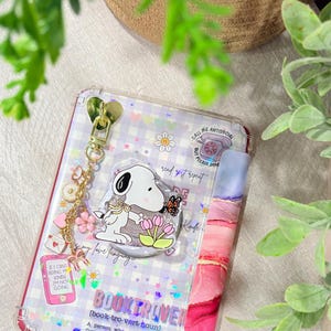 May include: A clear e-reader case featuring a purple and white checkered background with holographic stars. It includes a Snoopy character charm, a gold heart-shaped clip, and various stickers including a pink phone, a telephone, and the text BOOKTROVER.