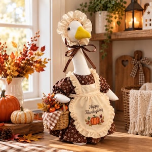 Puede incluir: Una figura de ganso blanco con un vestido floral marrón y un delantal de volantes color crema que muestra el texto Happy Thanksgiving y un gráfico de calabaza. El ganso lleva un gorro a juego y sostiene una pequeña cesta de mimbre.