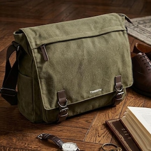 May include: An olive green canvas messenger bag featuring a fold-over flap with two brown faux leather straps, metal buckles, and a zippered pocket. The brand name TIMBRYN is printed in white on the front flap, and the bag has a dark brown strap.