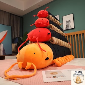 May include: A large, soft, orange and black plush centipede toy with multiple segments and legs, featuring long antennae and small black eyes. The toy has a bright orange head and body segments that transition into a long black tail section.