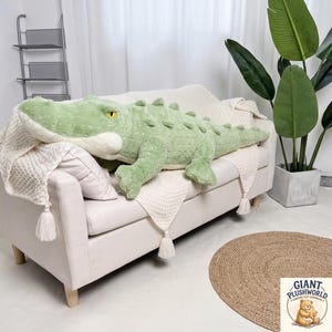 May include: A large, soft, light green plush crocodile toy with a textured, bumpy back, a cream-colored underbelly, and bright yellow eyes. The stuffed animal is positioned horizontally across a cream-colored sofa draped with a textured throw blanket.