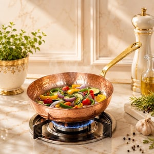 May include: A hammered copper skillet with a long gold-toned handle sits on a gas burner. The pan contains a colorful mixture of sautéed vegetables, including sliced red onions, yellow bell peppers, green zucchini, and bright red cherry tomatoes.