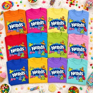 May include: A collection of twelve short-sleeved t-shirts in various solid colors, each featuring the Nerds candy logo with colorful cartoon characters printed on the front. The shirts are arranged in a grid pattern on a white surface.