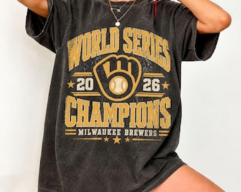 May include: A charcoal grey short-sleeve t-shirt featuring a distressed yellow graphic that reads WORLD SERIES 2026 CHAMPIONS MILWAUKEE BREWERS with a baseball glove logo in the centre and small stars flanking the text.