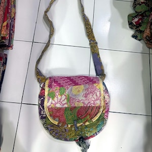 Pode incluir: Uma bolsa tiracolo com um design de retalhos de vários tecidos florais e estampados em tons de rosa, roxo, verde e amarelo. A bolsa tem uma aba arredondada, uma alça de ombro longa ajustável e um fecho de anel de metal circular.
