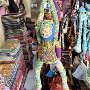 Puede incluir: Un peluche de mono colgante hecho de tela batik multicolor con una cara azul. El mono presenta brazos y piernas largos con intrincados patrones florales y geométricos en tonos verde, naranja, amarillo y morado, suspendido por un gancho de metal.