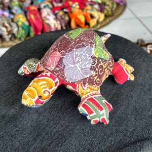 May include: A stuffed turtle toy crafted from multicolored fabric with a batik-style floral pattern on the shell. The head and limbs feature vibrant yellow, red, and green geometric and swirl designs, with small black beads serving as the eyes.