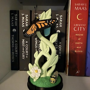 May include: A decorative figurine featuring a monarch butterfly perched atop a pale green plant stem with leaves. A small white flower and a striped caterpillar sit on the green base. A blue chrysalis hangs from the stem behind the butterfly.