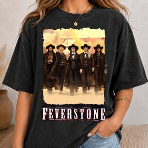 May include: A charcoal gray short-sleeve t-shirt featuring a graphic print of five individuals in western-style attire, including long coats and hats, standing in a desert landscape. The text FEVERSTONE is printed in a bold, distressed white font below.