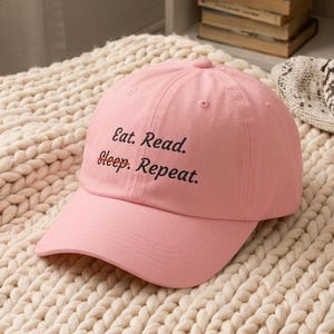 May include: A light pink baseball cap featuring the black embroidered text Eat. Read. Sleep. Repeat. on the front panel. The cap has a structured crown, a curved brim, and a button on top, constructed from a solid cotton fabric material.