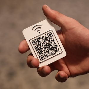 May include: A rectangular white plastic card featuring a raised black QR code in the center and a black Wi-Fi signal icon positioned above the code. The card has rounded corners and a smooth, matte finish, displaying a high-contrast graphic design.