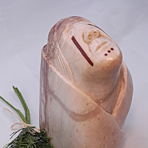 May include: A hand-carved stone sculpture of a person's head with a closed mouth and eyes, with red lines and dots painted on the face. The sculpture is made of a light brown stone with white and pink streaks.