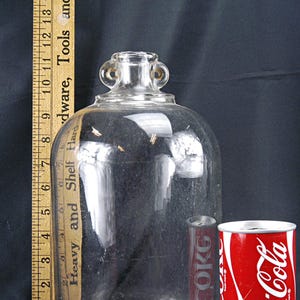 May include: A large, clear glass jug with a rounded body and two small handles near the neck. A Coca-Cola can is next to the jug for size comparison. A wooden ruler is on the left side.