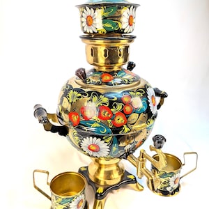 May include: A black and gold metal samovar featuring hand-painted floral motifs including white daisies and red berries, with a matching lid and two small metal cups with handles, all decorated with the same floral pattern.