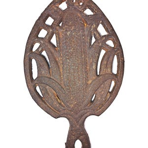 May include: A cast iron trivet with a rust-colored finish. The trivet has a stylized floral design with openwork details and a handle with a hole for hanging. The overall shape is reminiscent of a leaf.