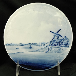 May include: A white ceramic plate with a blue and white windmill scene. The plate features a windmill, a house, and a river with a boat. The scene is painted in a traditional blue and white style.