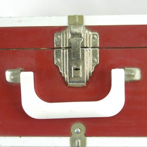 May include: Close-up of a vintage red suitcase with silver trim. The suitcase features a white handle and silver latches and a central locking mechanism. The case is likely made of a durable material, suggesting it was designed for travel.
