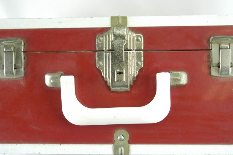 May include: Close-up of a vintage red suitcase with silver trim. The suitcase features a white handle and silver latches and a central locking mechanism. The case is likely made of a durable material, suggesting it was designed for travel.