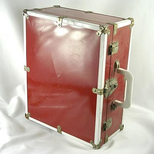May include: A vintage red suitcase with white trim and metal corner protectors. The suitcase has a white handle and metal latches. The exterior shows signs of wear, suggesting it has been used. This travel case is a classic piece.