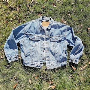May include: A light blue denim jacket featuring a classic collar, long sleeves with buttoned cuffs, two chest pockets with buttoned flaps, and a front button closure. A tan leather-like brand patch is visible on the inner neck area.