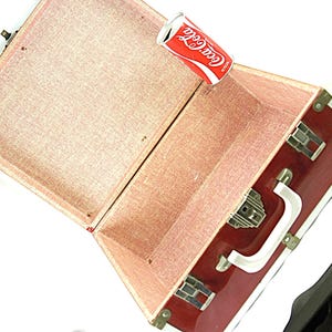 May include: Open vintage red suitcase with brass hardware and a white handle. The interior is lined with pink fabric. A Coca-Cola can rests inside the open suitcase, with the Coca-Cola logo visible.