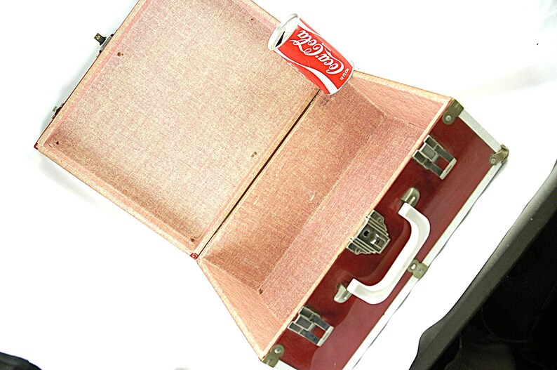 May include: Open vintage red suitcase with brass hardware and a white handle. The interior is lined with pink fabric. A Coca-Cola can rests inside the open suitcase, with the Coca-Cola logo visible.