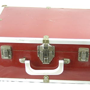 May include: A vintage red suitcase with silver trim and corner protectors. The suitcase has a white handle and a decorative silver clasp. The case is closed and appears to be made of a durable material.