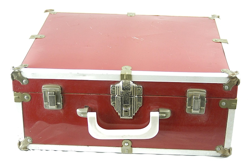 May include: A vintage red suitcase with silver trim and corner protectors. The suitcase has a white handle and a decorative silver clasp. The case is closed and appears to be made of a durable material.