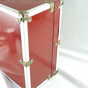 May include: A vintage red trunk with white trim and metal corner protectors. The trunk has a rectangular shape and is made of a sturdy material. The metal hardware is a gold color. The trunk is likely used for storage or travel.