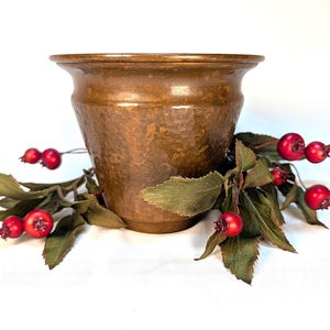 May include: A hammered copper-toned metal planter with a wide, flared rim. The vessel features a mottled, aged finish and is surrounded by a decorative sprig of dark green leaves and small, round, bright red berries.