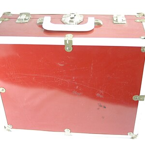 May include: A red vintage trunk with a white handle and trim. The trunk has metal corner protectors and clasps. The exterior shows signs of wear, with scratches visible on the surface. The trunk is designed for storage and travel.