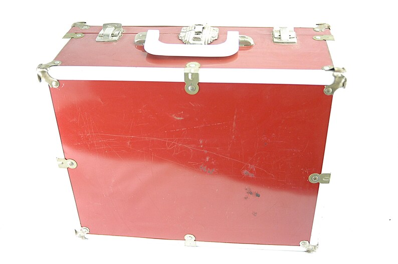May include: A red vintage trunk with a white handle and trim. The trunk has metal corner protectors and clasps. The exterior shows signs of wear, with scratches visible on the surface. The trunk is designed for storage and travel.