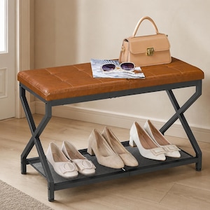 May include: A rectangular bench with a tufted tan leather-like seat and a black metal X-frame base. A lower mesh shelf holds four pairs of shoes. A beige handbag and a pair of sunglasses resting on a magazine are placed on the bench seat.