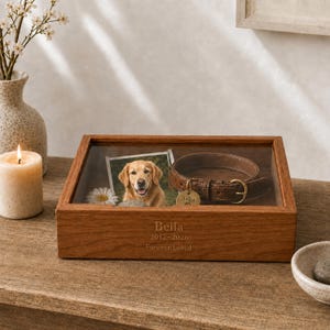 Puede incluir: Una caja de recuerdos rectangular de madera con tapa de cristal, grabada con el texto Bella, 2012-2026 y Forever Loved. Dentro hay una fotografía de un golden retriever, un collar de perro de cuero marrón con una placa y una flor blanca.