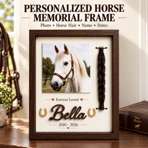 May include: A dark wood memorial shadow box frame featuring a white background, a square photograph of a white horse, a braided lock of dark horse hair, the name Bella in script, two small horseshoe icons, and the dates 2010-2026 in black text.